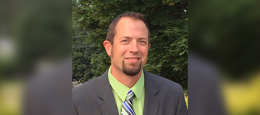 David Burke, Director of Operations at ACRT, wearing a gray suit with a green shirt and striped tie, standing outdoors in front of trees.