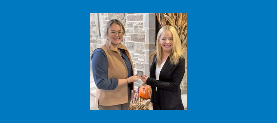 ACRT Operations Manager Anna Davis receives the ACRT Safety Challenge Coin from ACRT Safety Manager Adrienne Jones. Both are smiling while standing outside a stone building decorated with corn stalks.