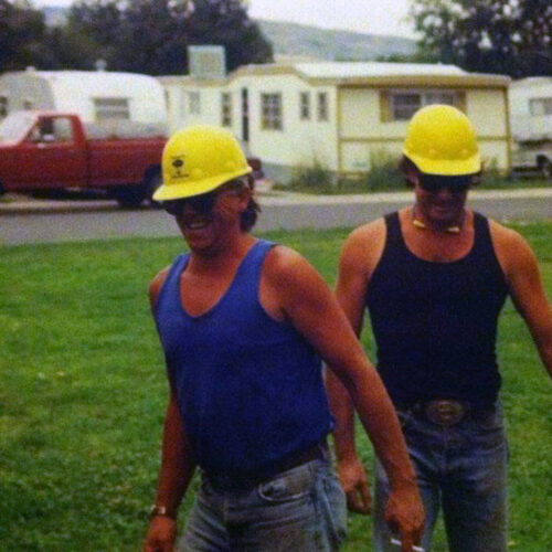 Ricky Slaven - Throwback Two workers wearing yellow hard hats and tank tops walk across a grassy area, with a red pickup truck and mobile homes in the background.