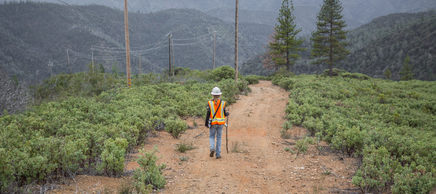 Utility worker in high-visibility safety gear walks along a dirt access road beneath power lines in a mountainous landscape, demonstrating field awareness and environmental stewardship.